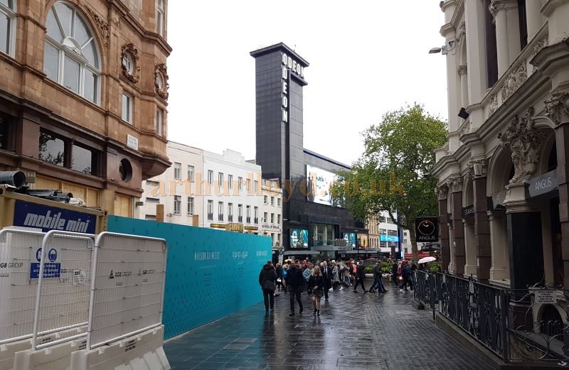 Leicester Square from Leicester Place in April 2017, showing the site of the former Sans Souci Theatre to the near left. The photograph also shows the Odeon, Leicester Square which was built on the site of the former Royal Panopticon of Science and Art, and the Alhambra Theatre.